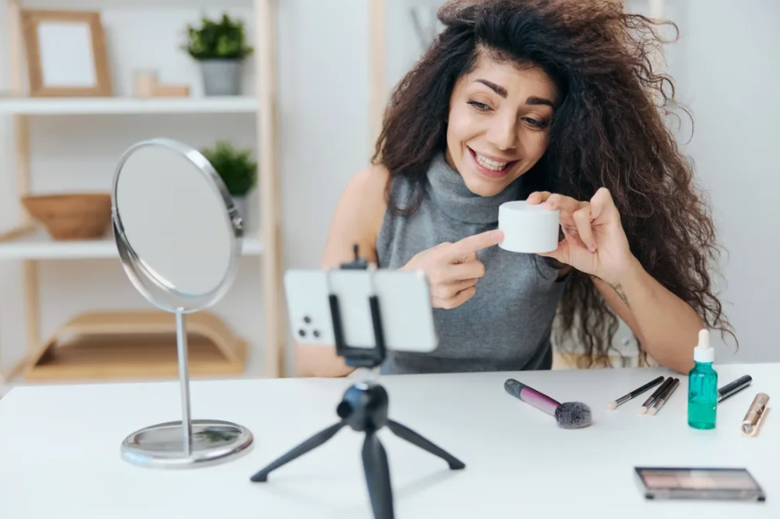 Woman filming a makeup tutorial for an online business, showcasing beauty products on a smartphone tripod.